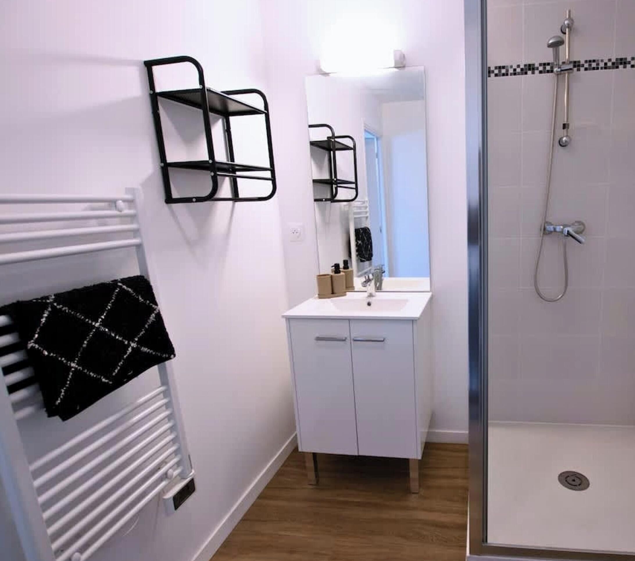 A bathroom with a white sink, mirror, and shower. There are black towel racks on the wall and a white radiator.