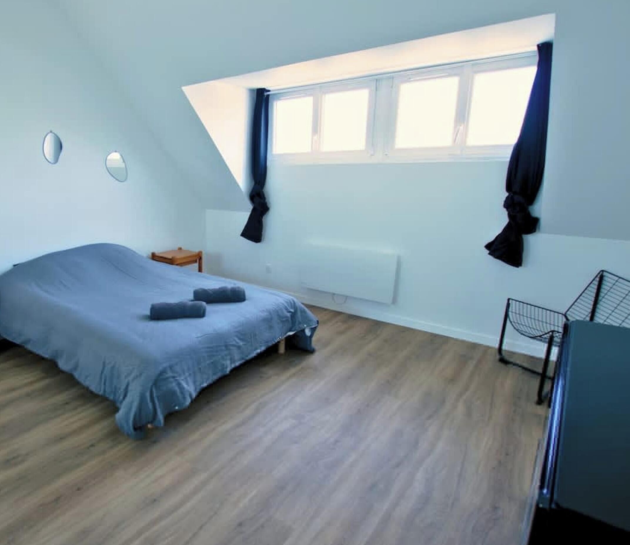 A bedroom with a sloped ceiling, a bed, a small wooden nightstand, a radiator, and a window with curtains.