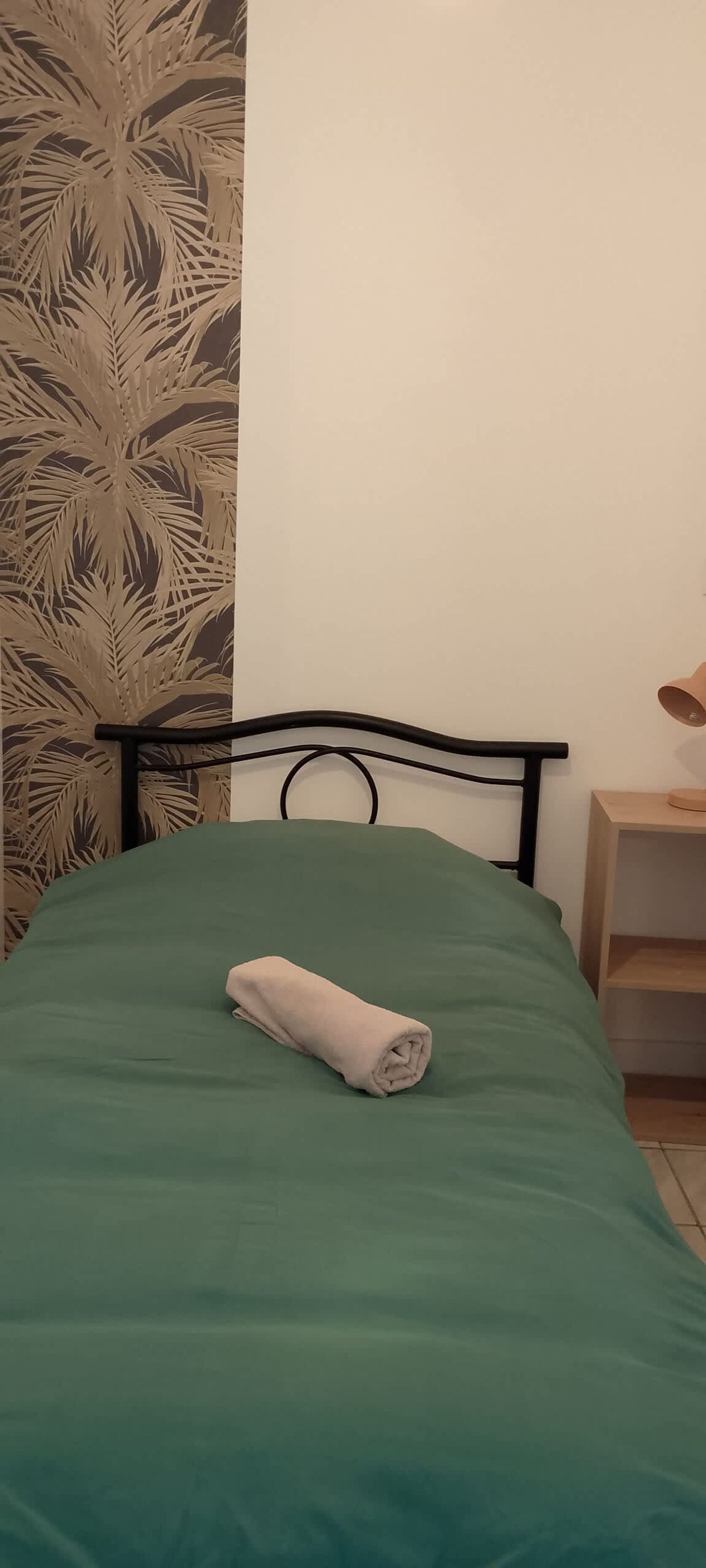 A bed with a green bedspread and a rolled towel, a black metal headboard, a wooden nightstand with a lamp, and a wall with a palm leaf pattern.