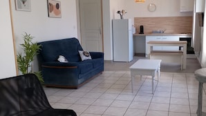A living room with a sofa, a coffee table, a dining table, and a kitchen area with cabinets and appliances. - Chez Serge N 107 Jardin Parking Animaux 4 Pers (Noeux-les-Mines)