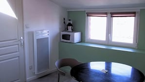 Une pièce avec une porte blanche, un réfrigérateur blanc, un four à micro-ondes, une table avec une chaise et une fenêtre avec des stores.