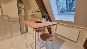Una pequeña mesa con marco blanco, superficie de madera y dos taburetes, una planta en maceta y una ventana con vistas a árboles desnudos.