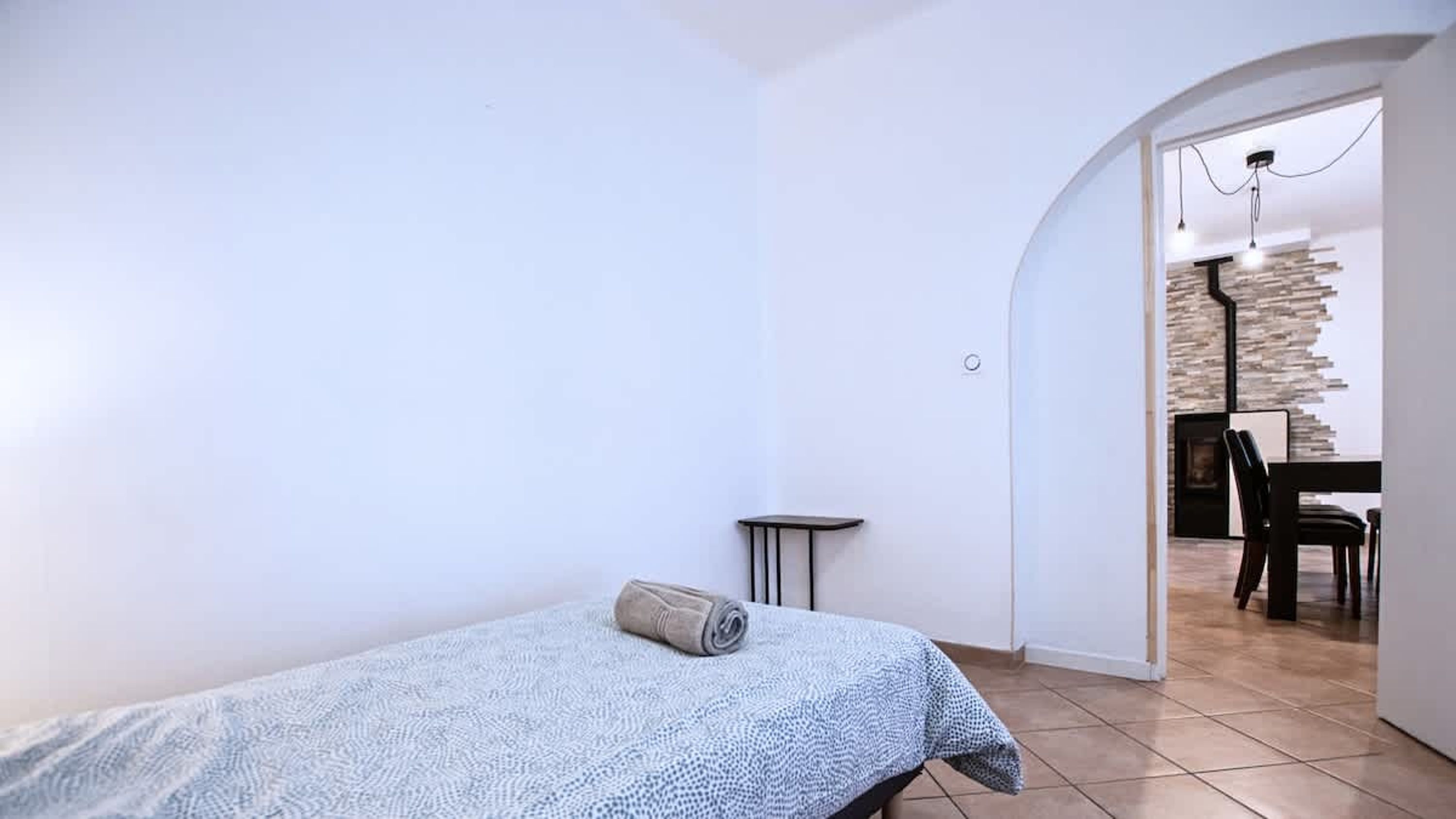 A small, white-walled room with a bed, a small table, and a doorway leading to a dining area with a stone wall and a table.