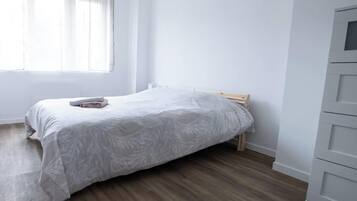 A bedroom with a bed, wooden floor, white walls, and a window with sheer curtains.