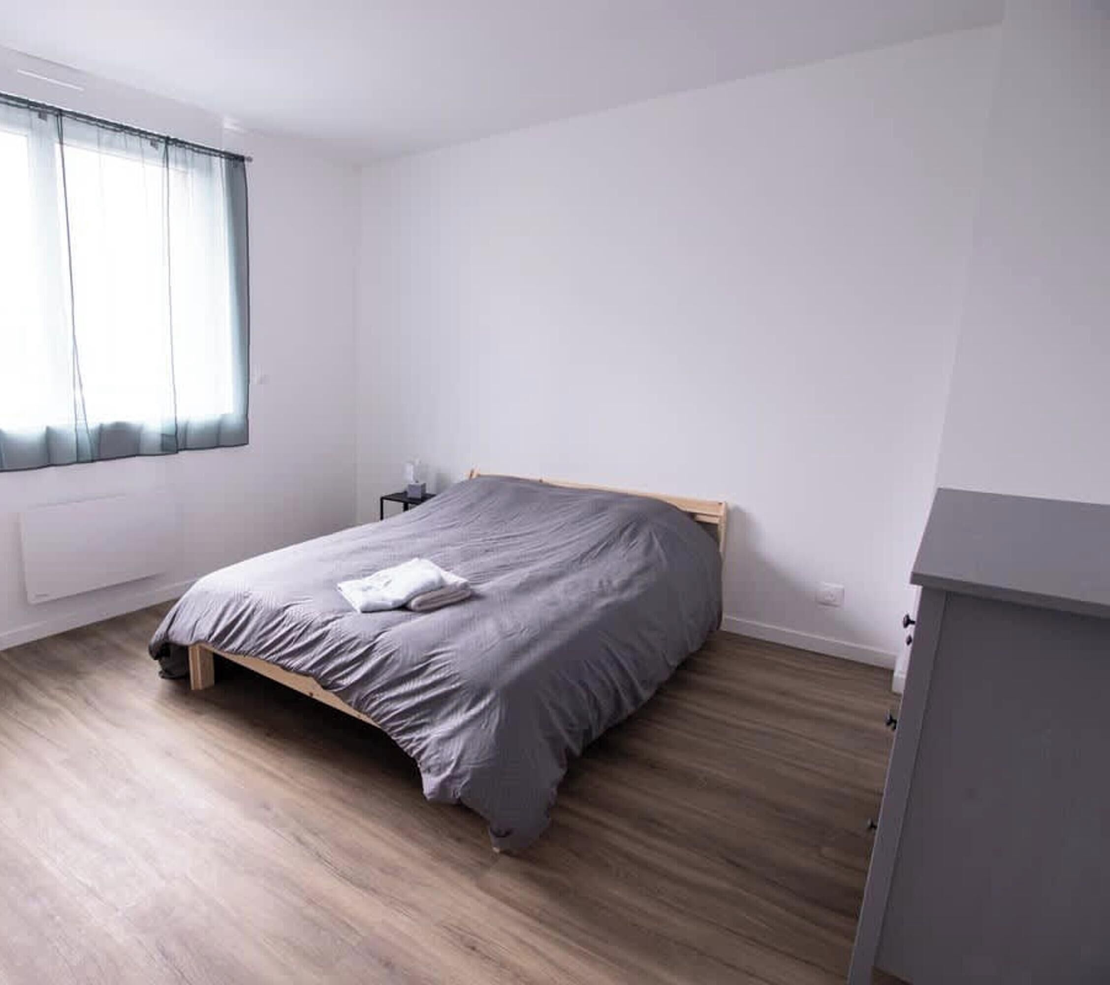 A bedroom with a bed, wooden flooring, a nightstand, a window with curtains, and a small desk.