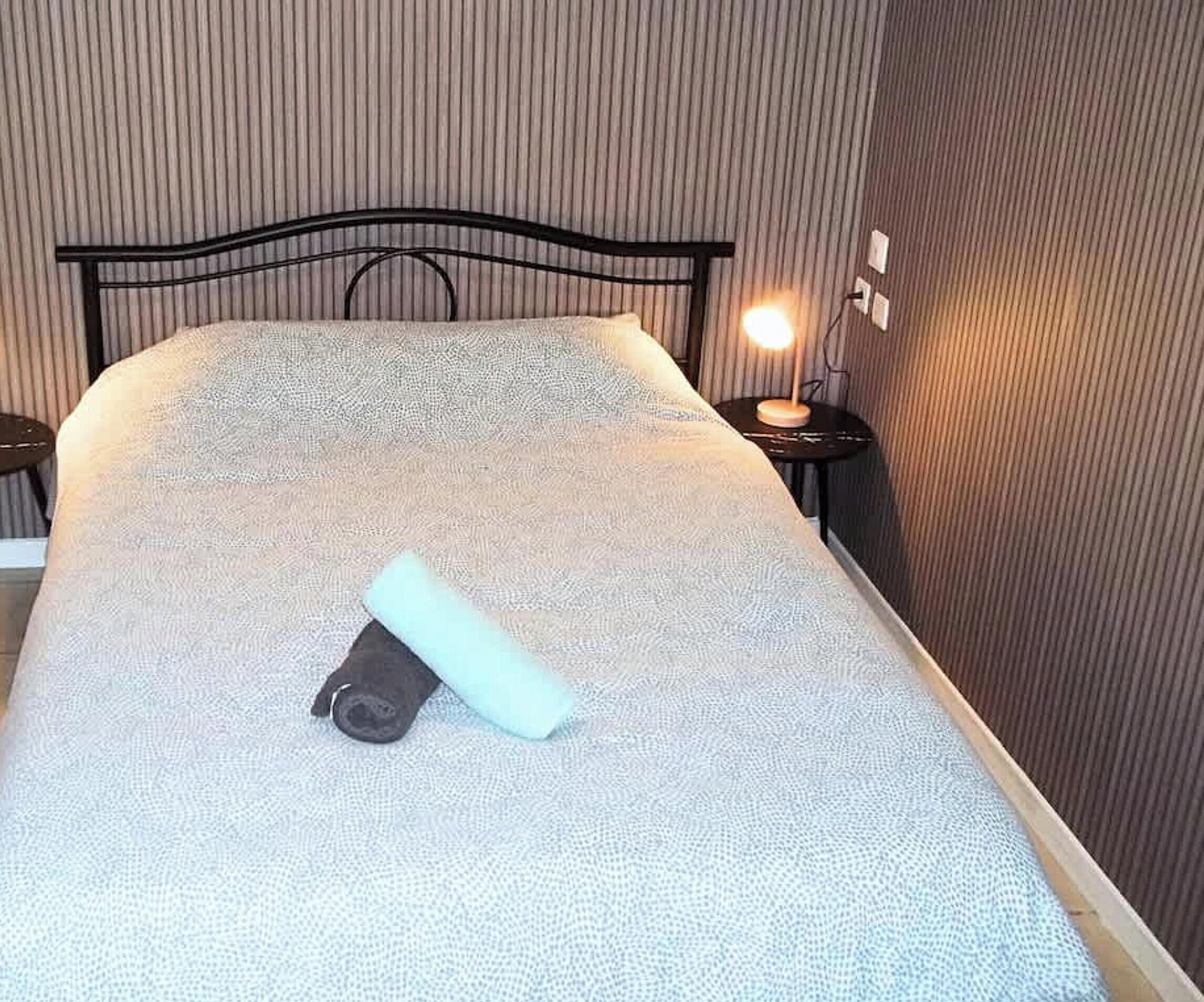 A neatly made bed with a patterned bedspread, a rolled-up towel, and a bedside lamp on a nightstand.