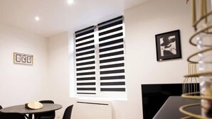 Une pièce avec une porte noire, une table ronde entourée de chaises, une fenêtre avec des stores et un tableau encadré accroché au mur.