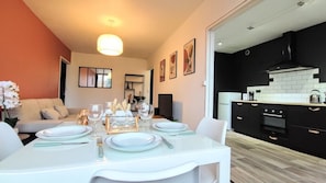 A dining area with a white table set for four, a sofa in the background, and a kitchen with black cabinets and white tiles. - Atelier Wagram Au Coeur de Troucoing (Tourcoing)
