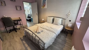 A bedroom with a bed, a desk, a chair, a bedside table, and a wall-mounted artwork.