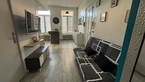 A compact living space with a sofa, a TV mounted on the wall, a small kitchen area, and a window with blinds.