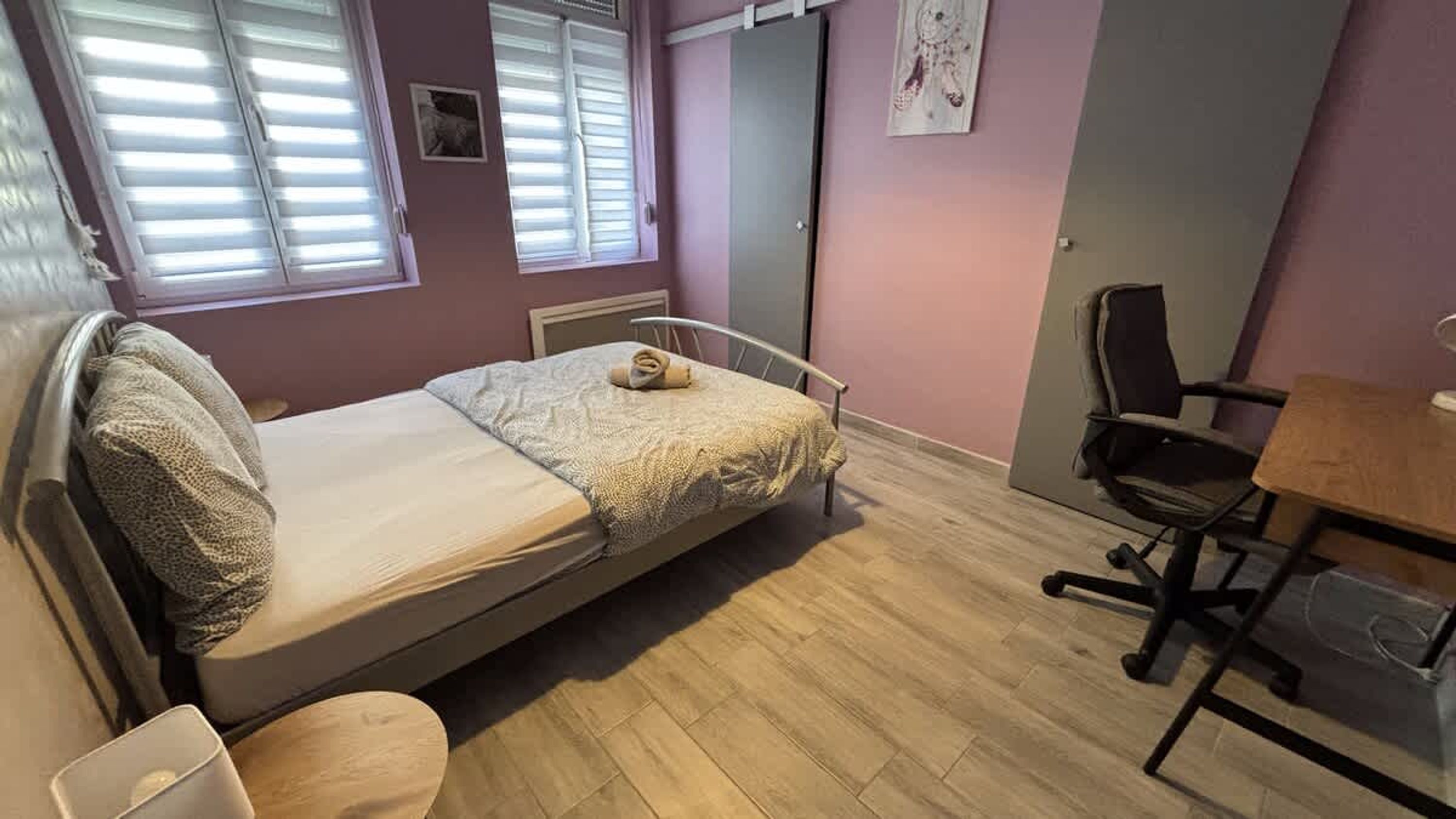 A single bed with a rolled towel, a desk, a chair, and a window with blinds.
