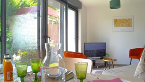 A modern room with a large window, a television, and a dining area with a table set for breakfast. - Le Matisse - Centre - Jardin au Calme - Lumineux (Béthune)