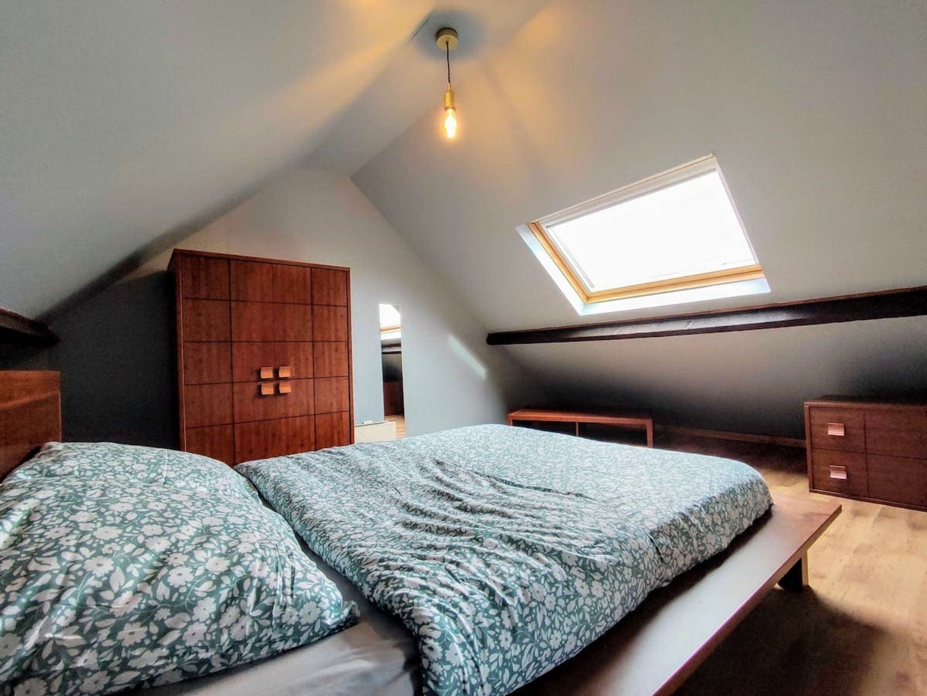 A bedroom with a skylight, a bed with floral bedding, a wooden wardrobe, and a bedside table.