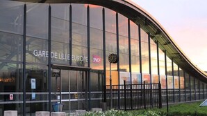 Um edifício moderno com grandes janelas de vidro, um relógio e placas com a inscrição 'GARE DE LILLE EUROPE' e 'SNCF'.