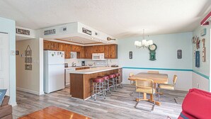 Una cocina compacta con armarios de madera, una isla central con taburetes, un comedor con mesa y sillas, y una sala de estar con sofá.