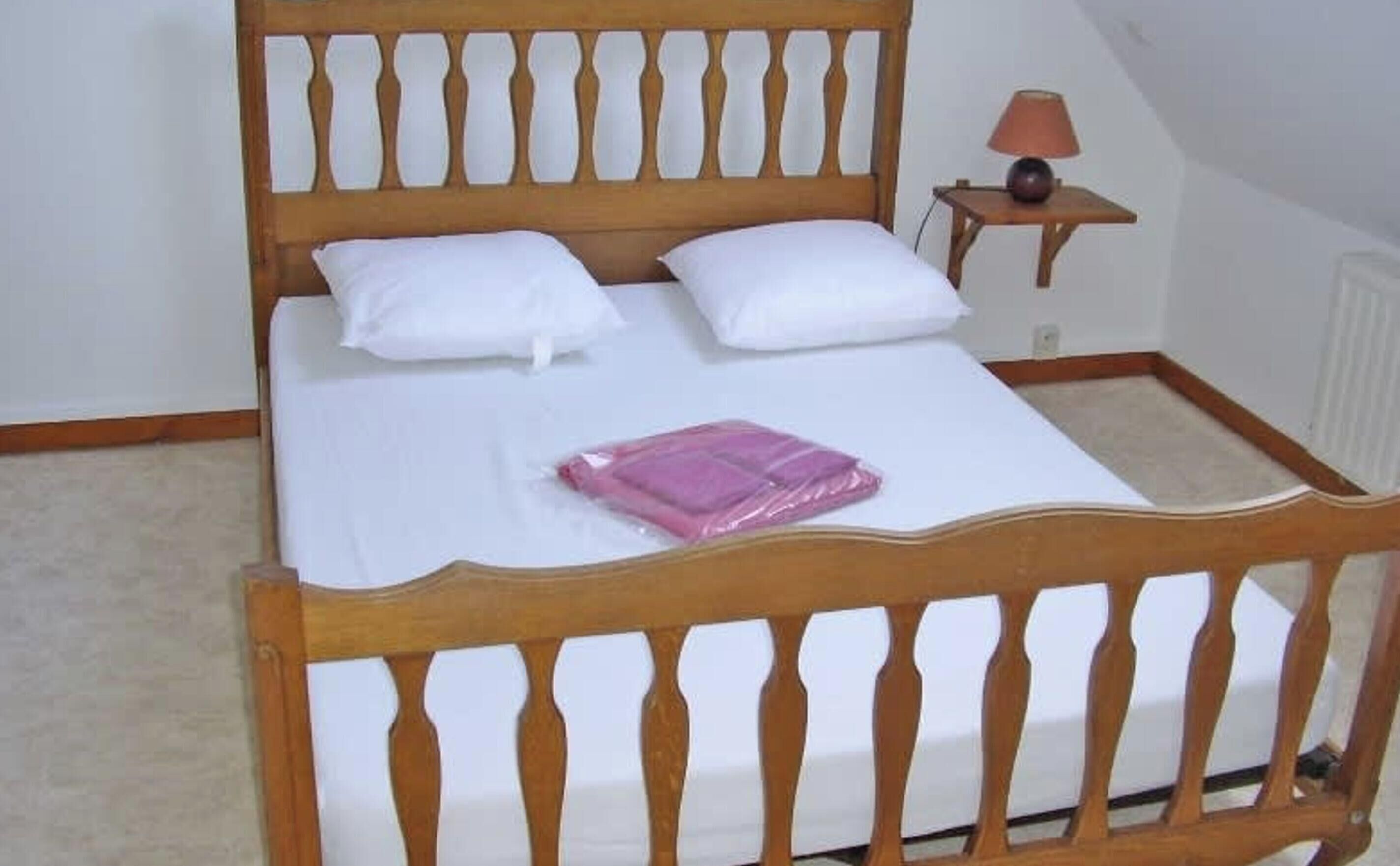 Un lit en bois avec une literie blanche, une petite table de chevet avec une lampe, et un paquet rose posé sur le lit.