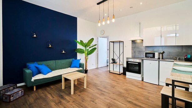 Una sala de estar moderna con un sofá, una mesa de centro y una planta grande. Junto a ella, una cocina con armarios blancos, un microondas y una estufa.