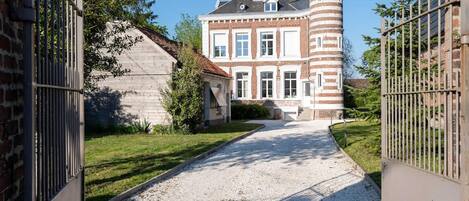 A gated driveway leading to a large, historic building with a prominent tower and a smaller attached structure.