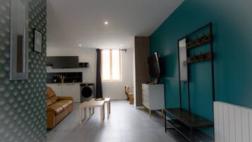 A modern living room with a sofa, coffee table, and a TV. There is a kitchen area with a washing machine and a dining table.
