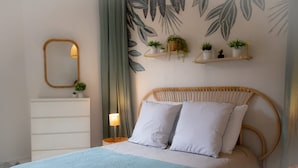 A bed with a woven headboard, white pillows, and a light blue blanket. There is a bedside table with a lamp and a mirror. Above the bed, there are wall shelves with plants. - Sublime 2 Bollaert 4 Pers Centre Ville (Lens)