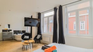 Ein modernes Hotelzimmer mit Bett, Fernseher, kleinem Tisch, zwei Stühlen, einer Couch und einem Fenster mit Vorhängen.