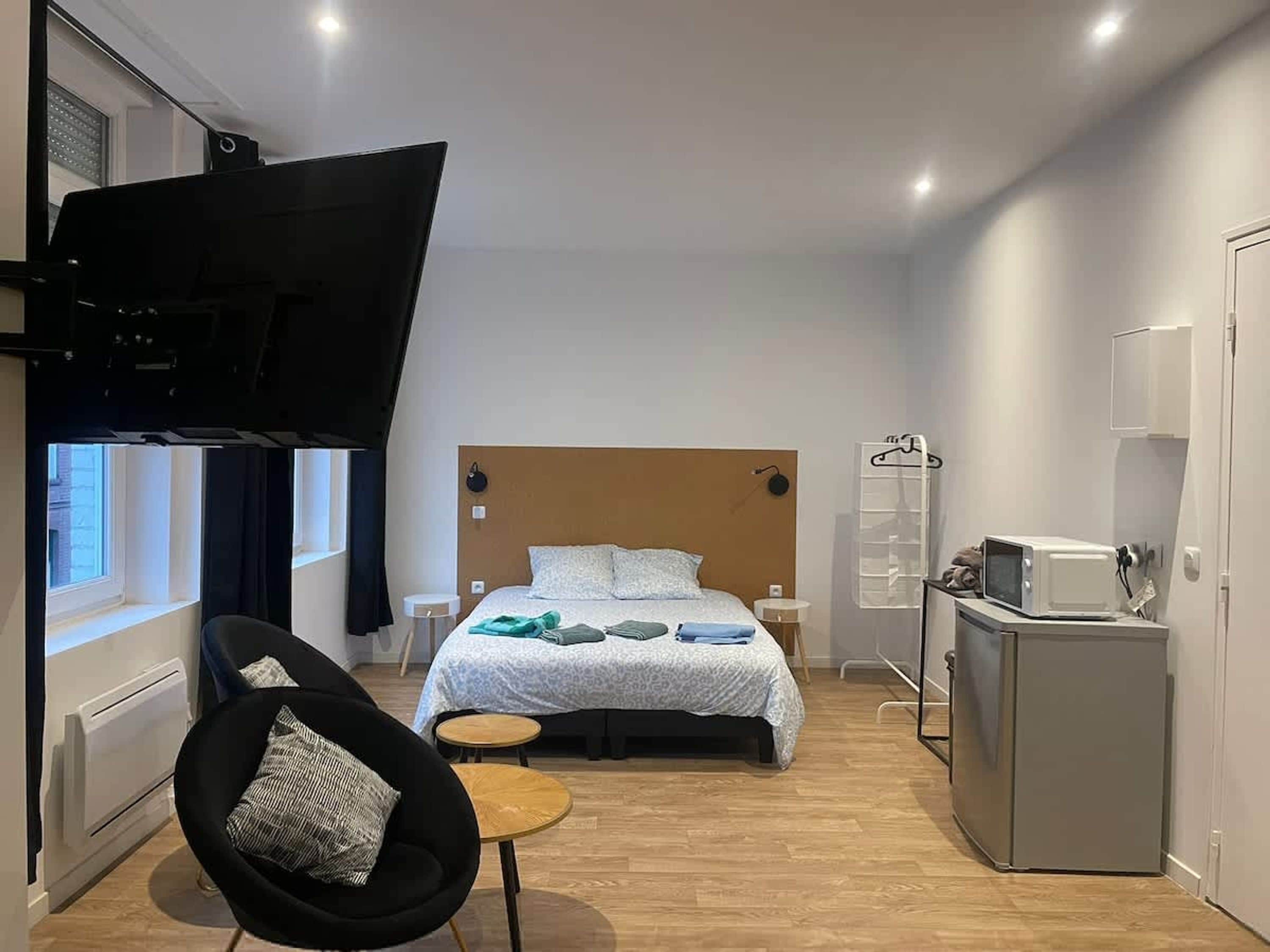 A compact hotel room with a bed, a small table, a chair, a TV, and a kitchenette.