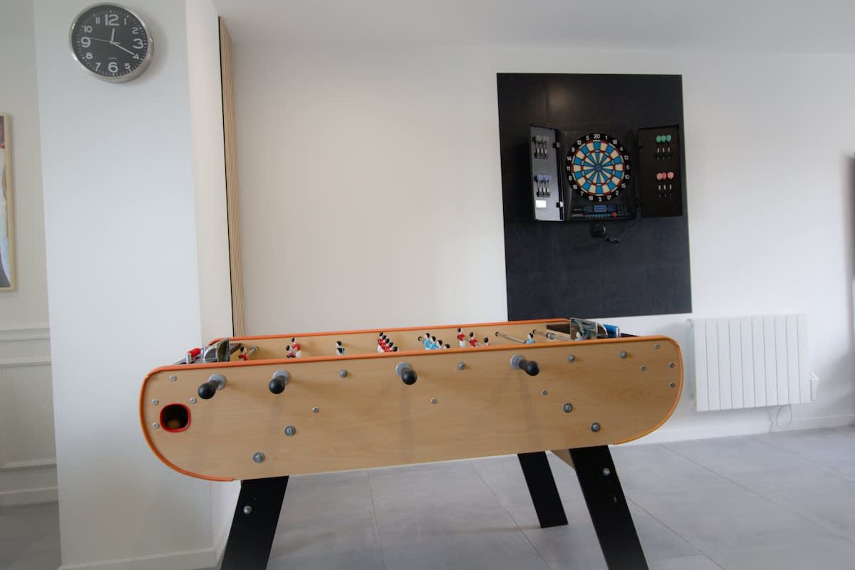 A foosball table, a dartboard, and a clock on the wall in a room with a tiled floor.