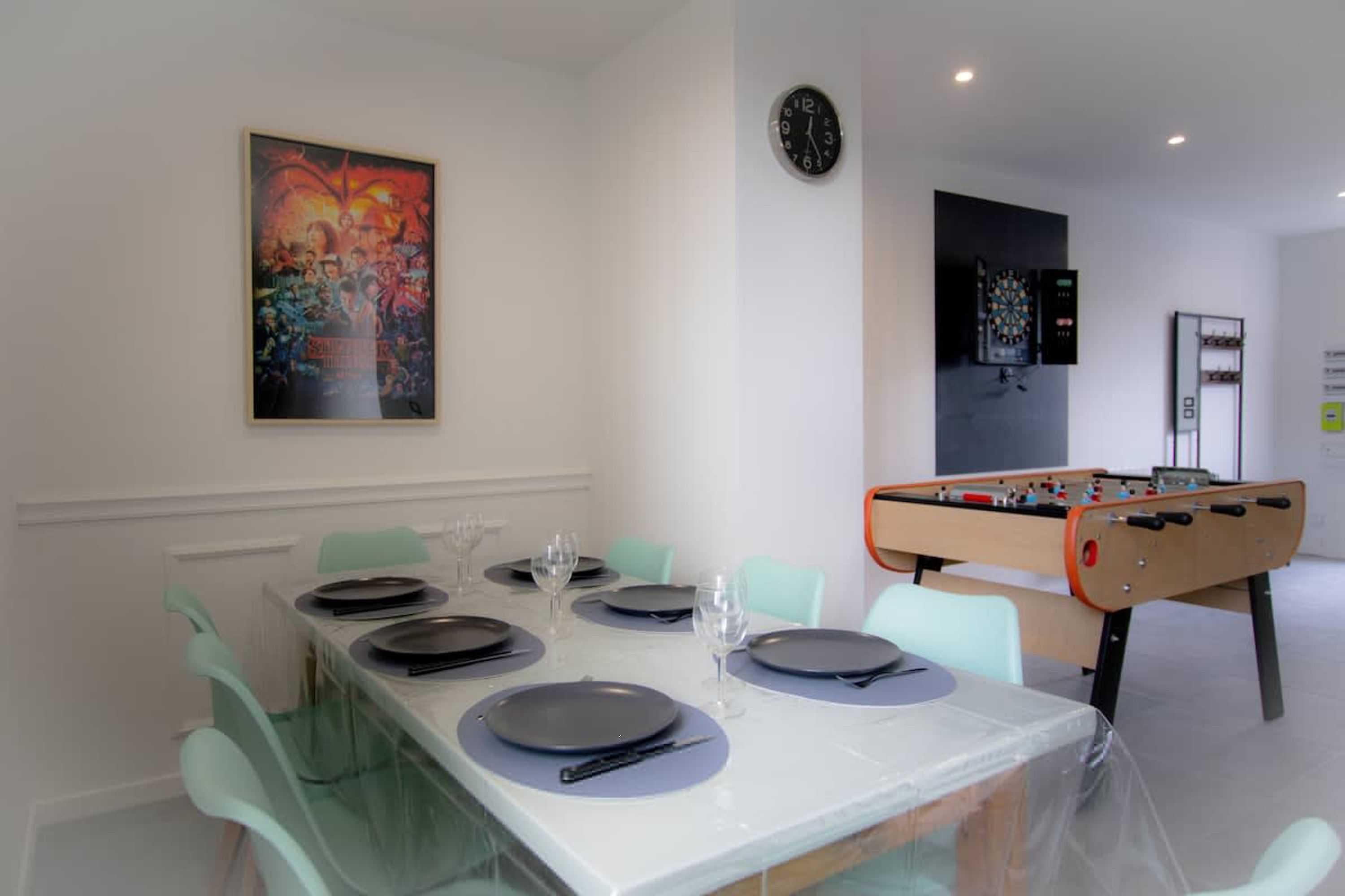 Un comedor con una mesa dispuesta para cuatro personas, mesa de fútbol de mesa, dardo y un reloj en la pared.