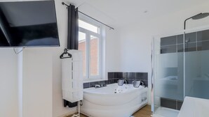 Une salle de bain moderne dotée d’une grande baignoire, d’un espace douche, d’un téléviseur fixé au mur et d’une fenêtre laissant entrer la lumière naturelle.