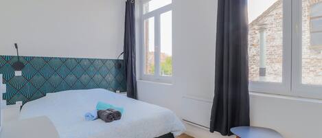 Un dormitorio con cama, ventana, cortina, una silla y vista a un edificio de ladrillo.