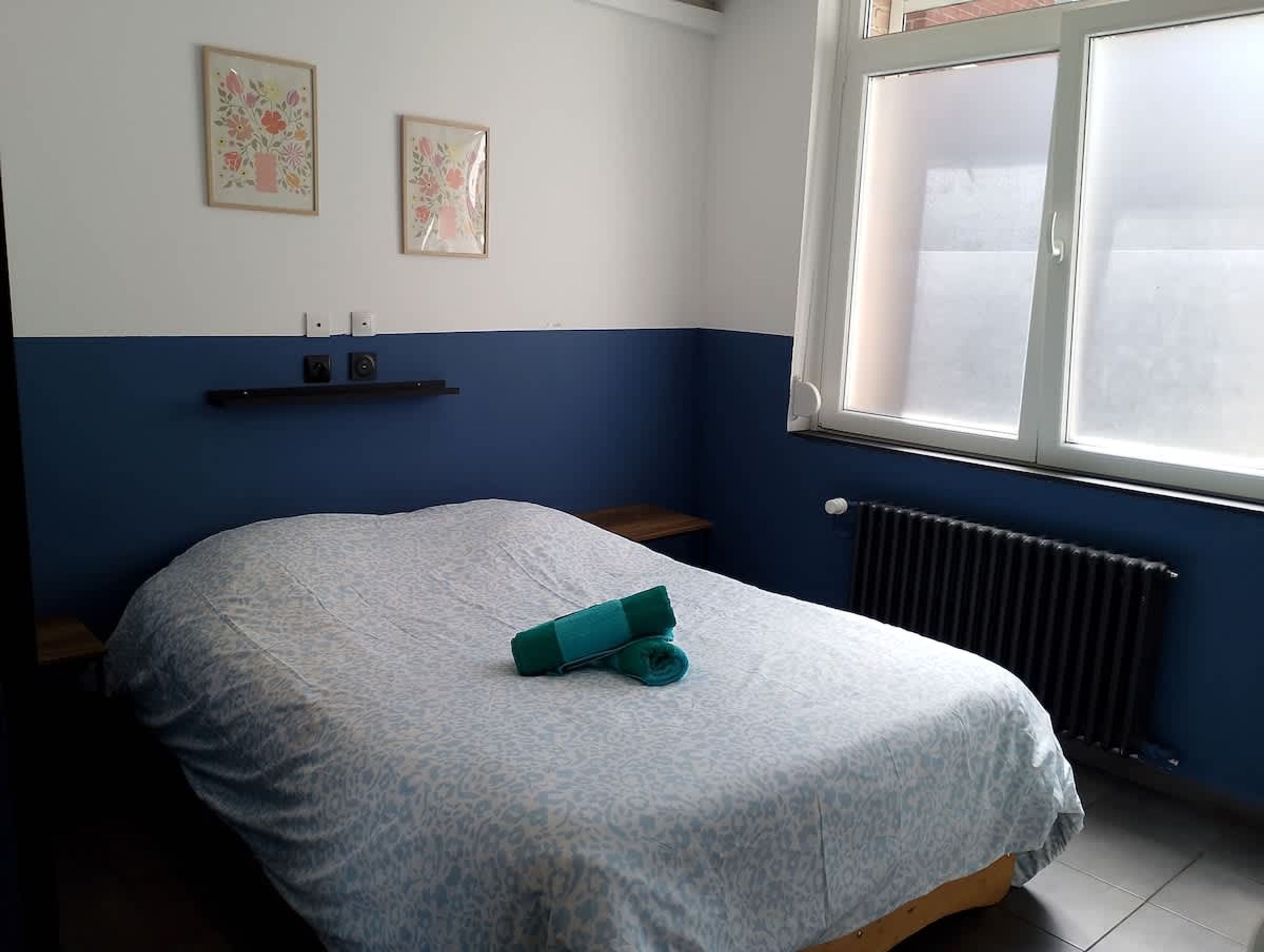 A bedroom with a bed, a radiator, a window, and two wall-mounted picture frames.