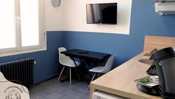 Una habitación pequeña con cama, escritorio, silla, televisor, radiador y ventana.