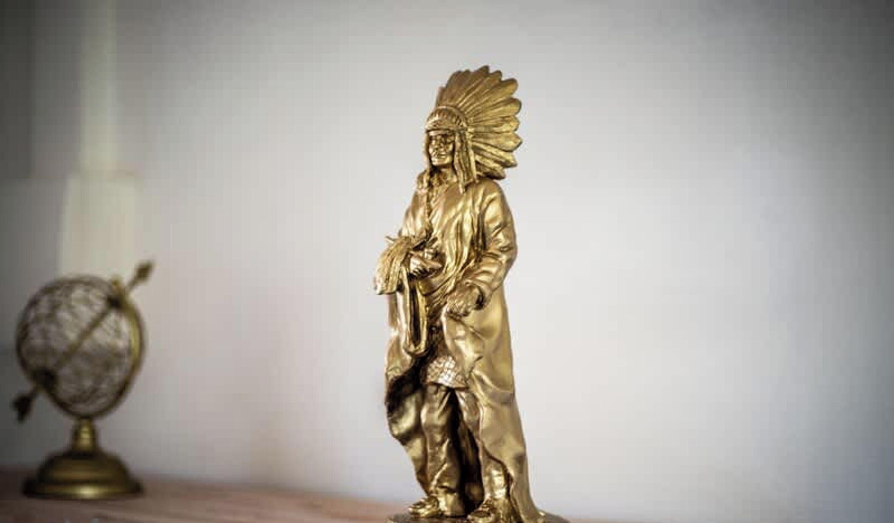 En gylden statue af en figur med en fjerprydet hovedbeklædning, der står på en overflade.