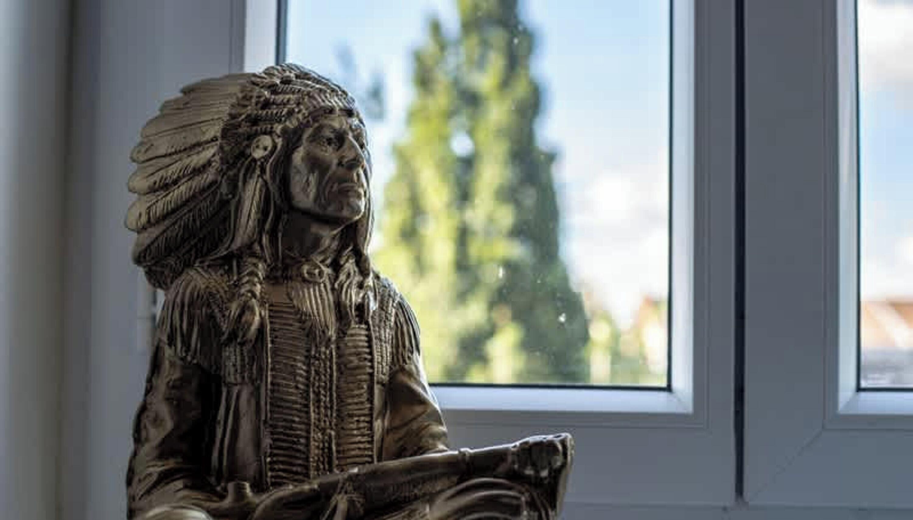 A statue of a person with elaborate headgear, positioned in front of a window with a view of trees and a building.
