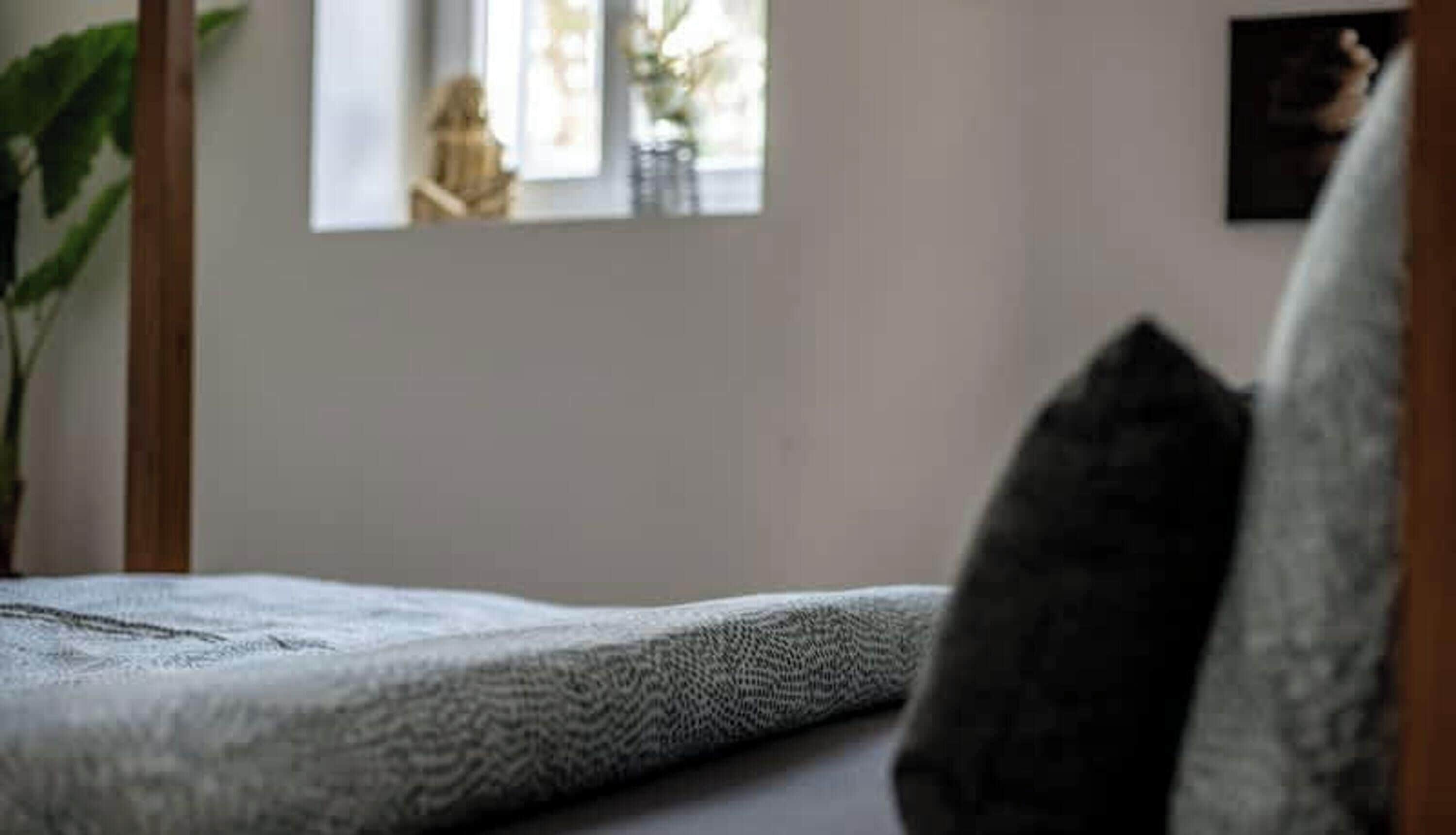 Een bed met een geribbelde deken, een donker kussen en een raam met uitzicht op iemand die buiten zit.