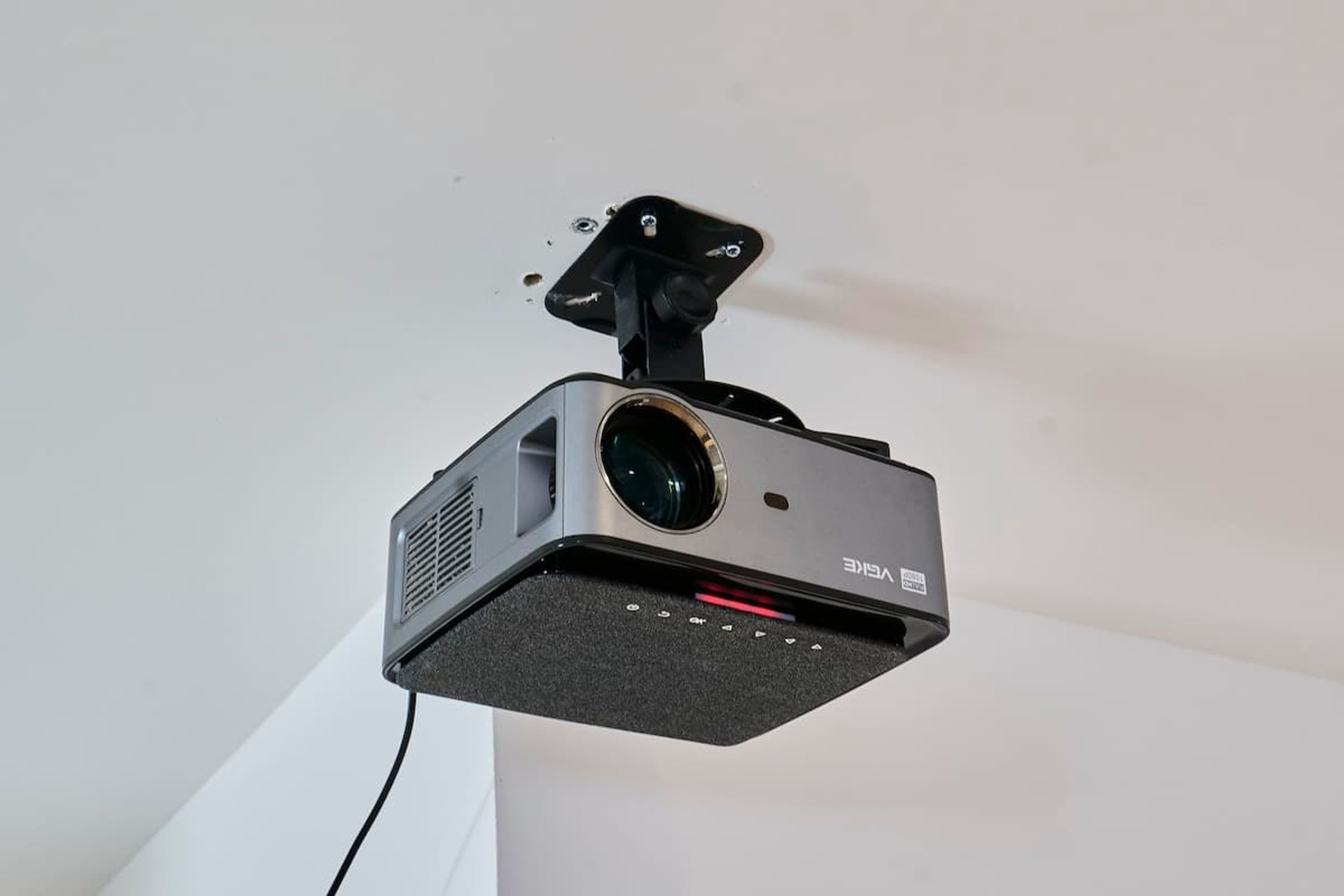 A ceiling-mounted projector with a lens, a control panel, and a mounting bracket.