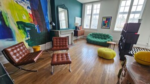 Ein Zimmer mit Kamin, einem grünen Sofa, einem gelben Pouf und einem großen abstrakten Gemälde.