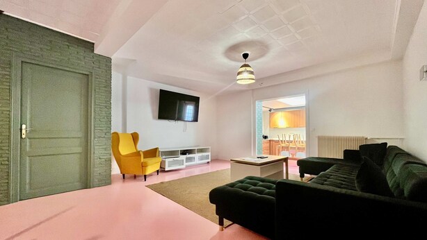 A modern living room with a sofa, armchair, TV, and a door leading to a kitchen. - Grande Maison 17 Personnes (Arras)