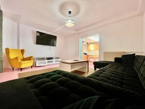 A living room with a sofa, a yellow armchair, a TV, and a dining area visible through an open door. - Grande Maison 17 Personnes (Arras)