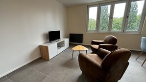 Ett vardagsrum med en tv, ett litet träbord, två bruna läderfåtöljer, ett fönster med utsikt över träd och ett vitt skåp.