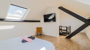 A bedroom with a skylight, a bed with rolled-up blankets, a chair, a TV, and a wooden ladder.