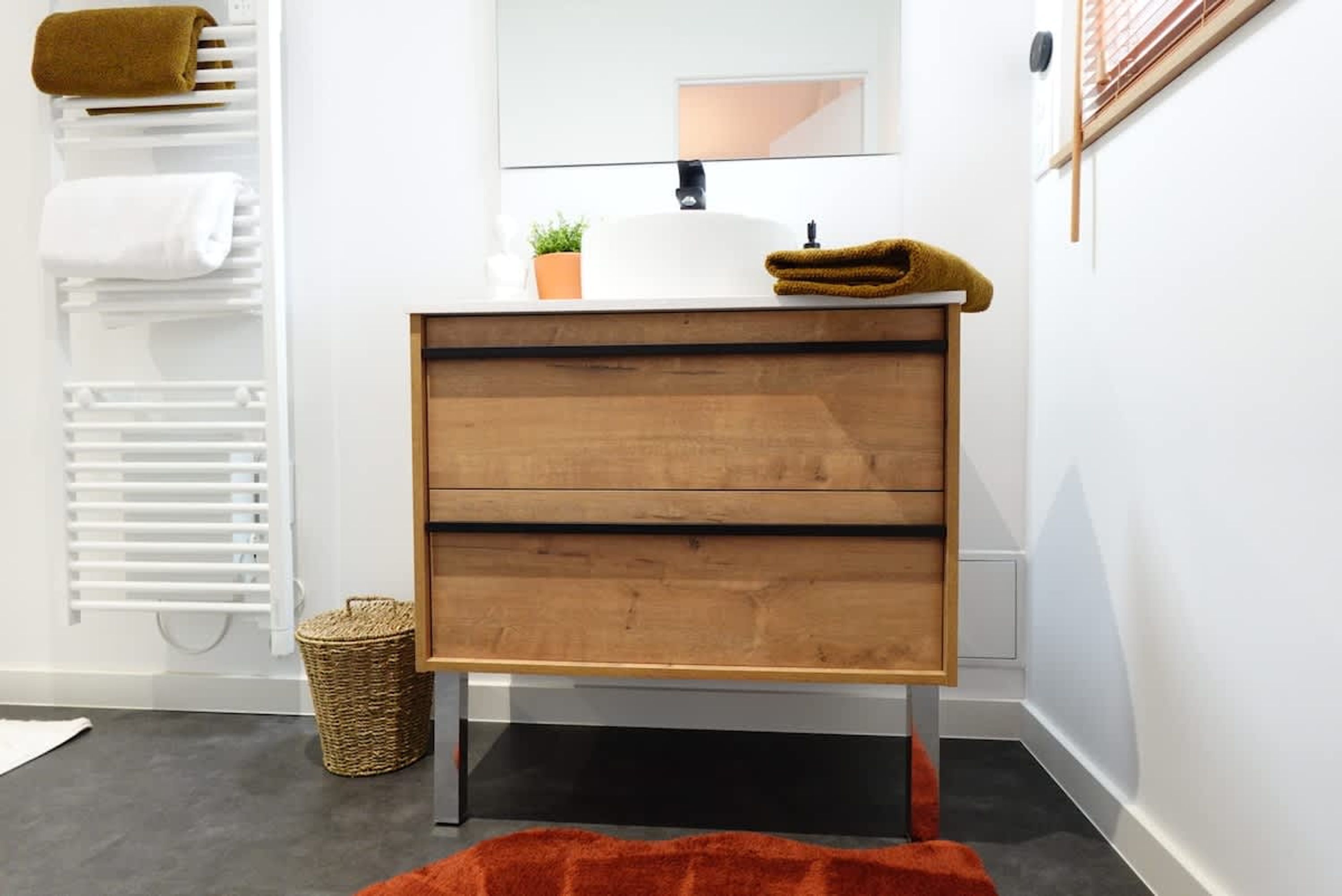 Ein Badezimmer mit einem weißen Waschbecken, einem Holz-Waschtisch, einem Spiegel, einem Handtuchhalter, einer Topfpflanze und einem Korbwäscheschrank.
