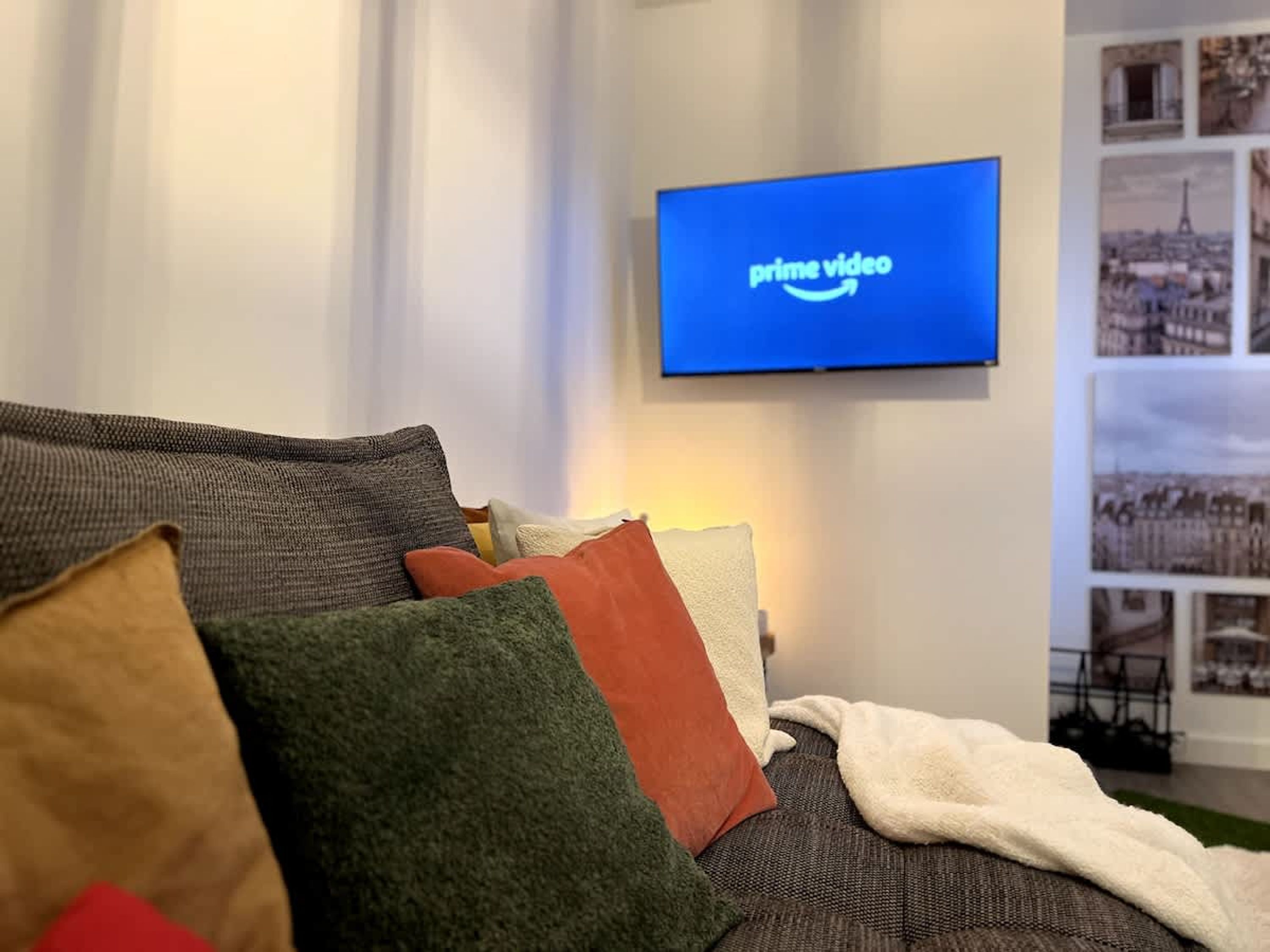 A living room with a flat-screen TV displaying the Prime Video logo, a sofa with multiple cushions, and a wall with framed pictures.