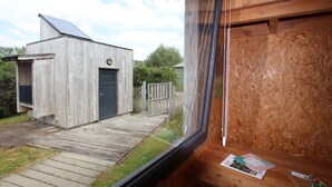 Een houten tuinhuis met een deur en een klein bijgebouw, een houten terras en een uitzicht op een huis door een raam.