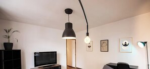 A modern living room with a flat-screen TV, a black pendant light, a potted plant, and framed artwork on the wall. - Le Voyageur Gare de Lens Wifi 4 Pers (Lens)