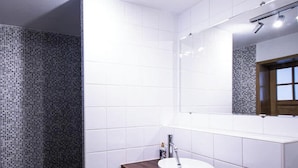 A bathroom with white tiled walls, a wooden vanity, a white sink, and a shower area with patterned tiles. - Le Voyageur Gare de Lens Wifi 4 Pers (Lens)