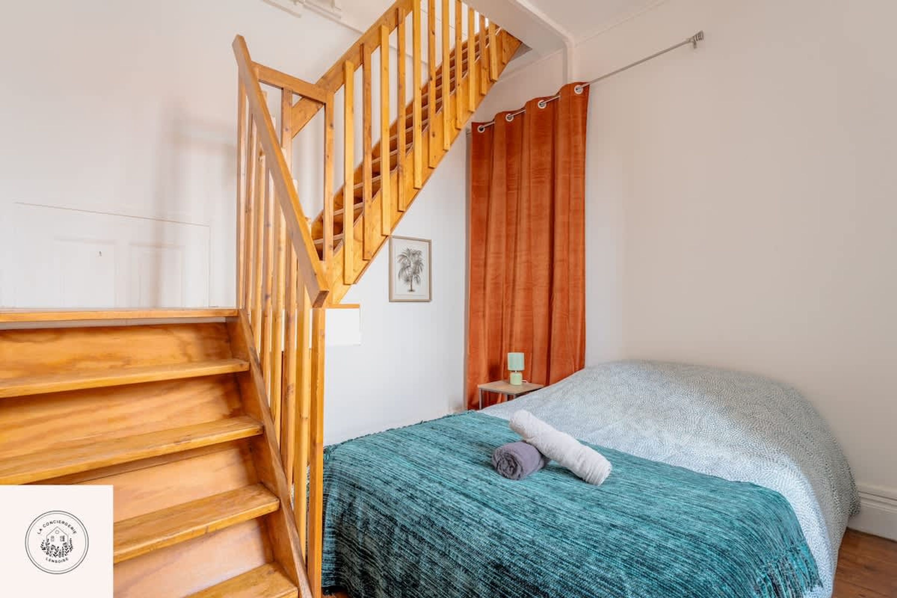 Une chambre avec un escalier en bois, un lit recouvert d’une couverture turquoise, une table de chevet avec une lampe et une tête de lit en bois.