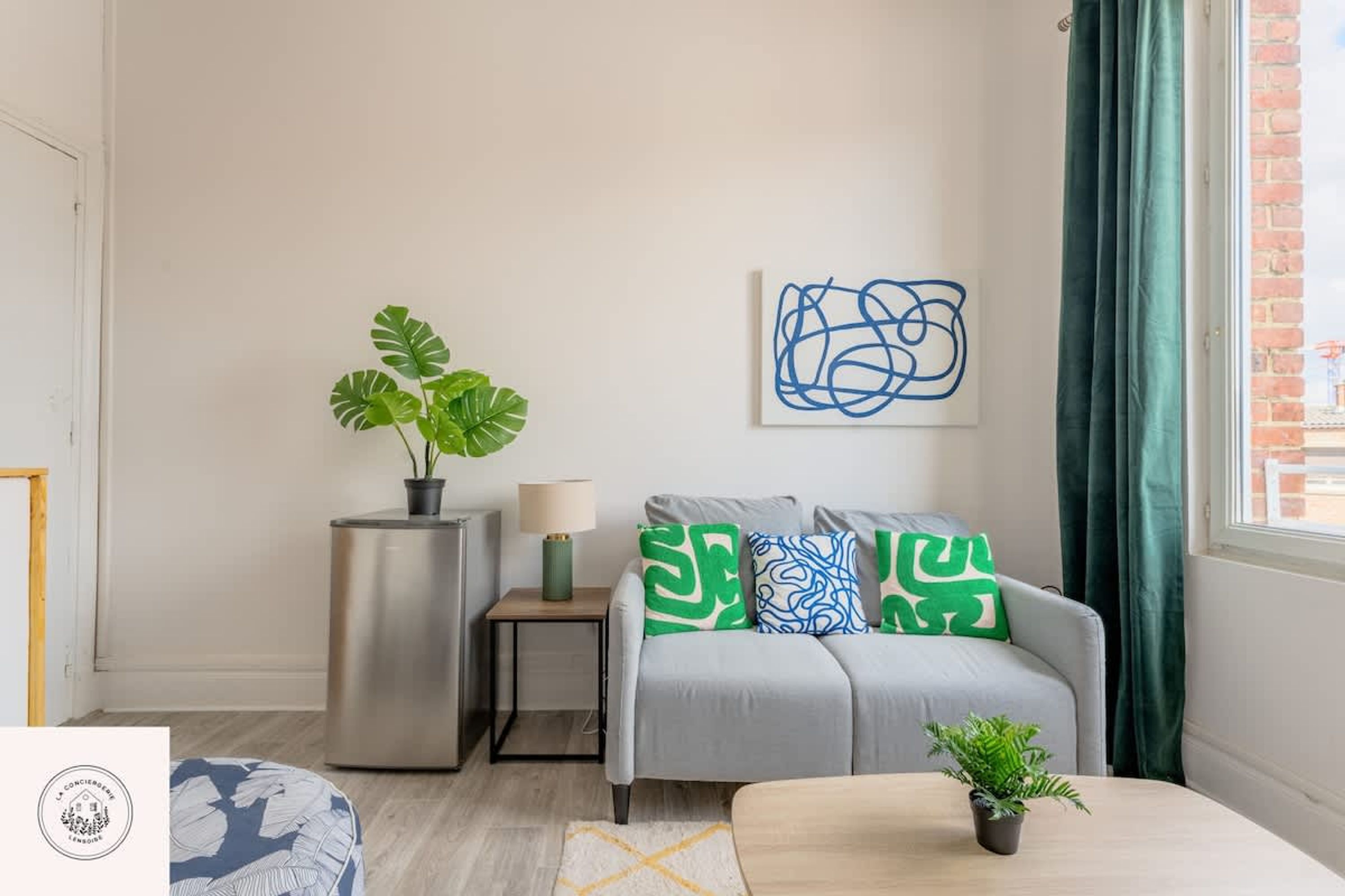 Una sala moderna con un sofá gris, cojines estampados en verde y azul, una planta en maceta y una obra de arte en la pared.