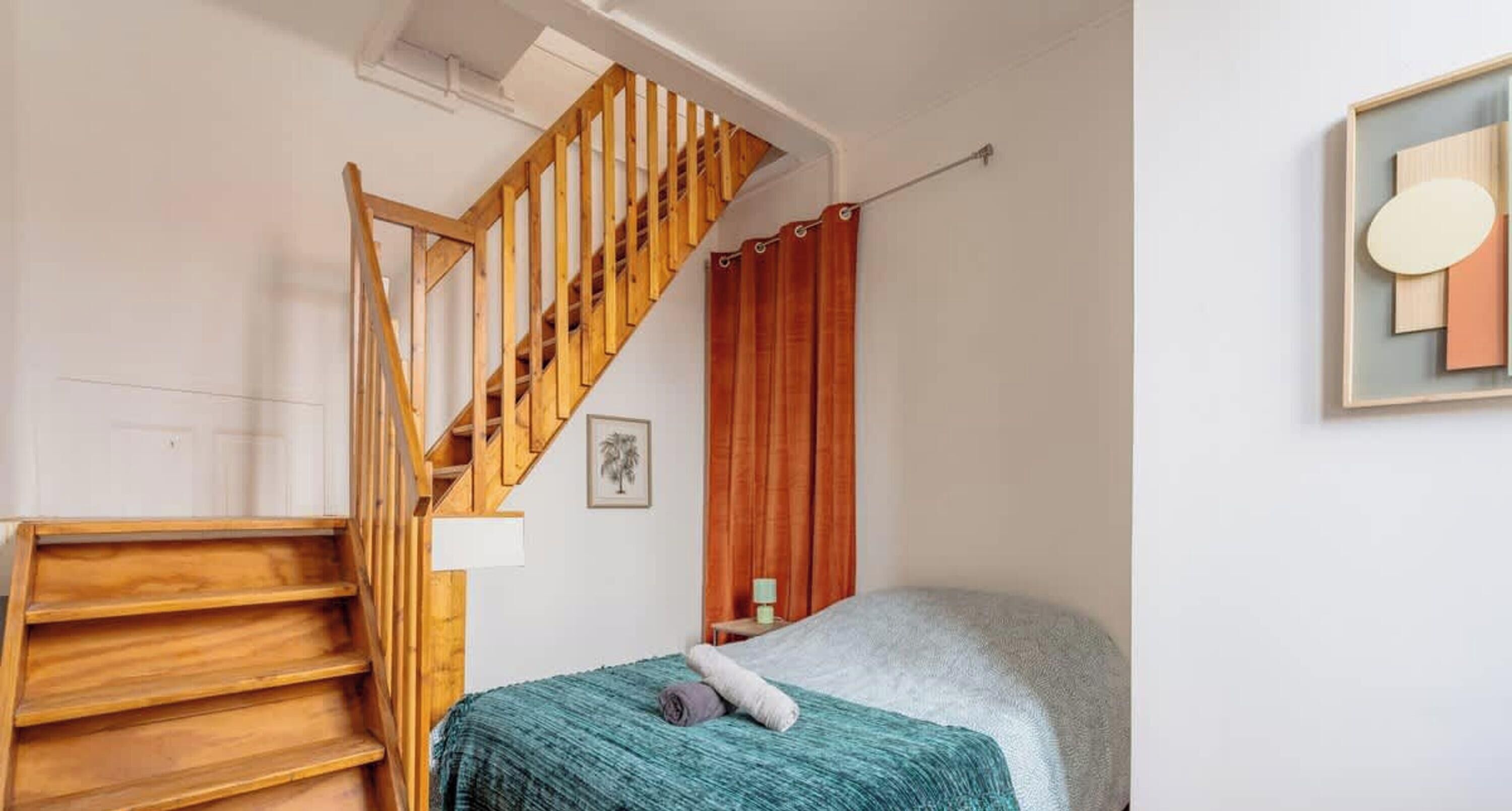 A bedroom with a wooden staircase, a bed with a teal blanket, a nightstand, and a wall-mounted artwork.