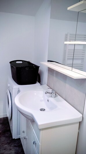 Uma casa de banho moderna com lavatório branco, cesto de roupa preto, máquina de lavar e espelho com prateleira.